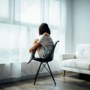A woman sitting on a modern black chair near a large window with sheer white curtains, gazing outside, symbolizing introspection, mood, or mental health